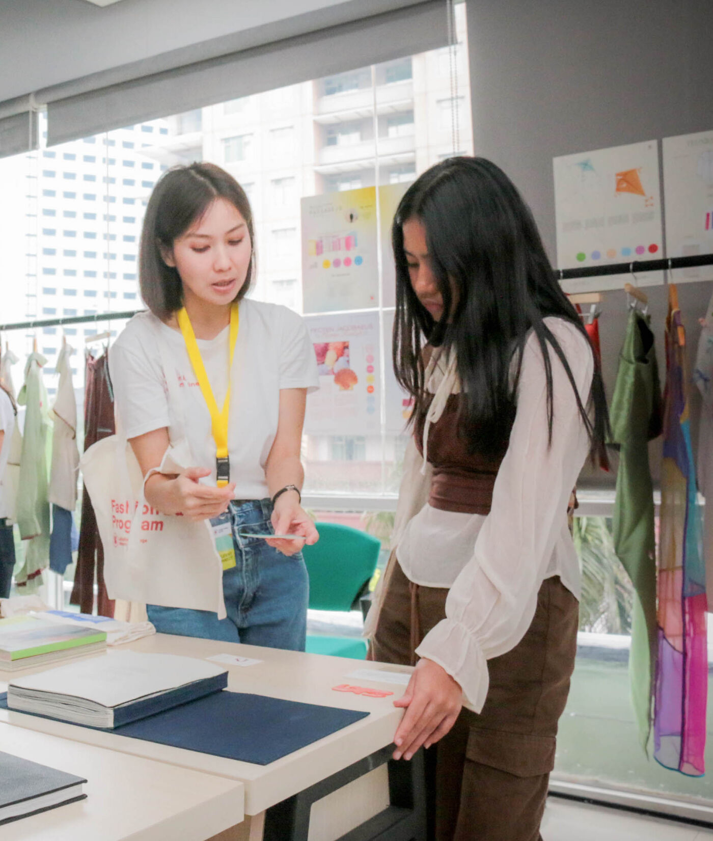 Two students engage in a discussion over design materials and portfolios during an open house at LCI Jakarta. Surrounded by fashion sketches, fabric samples, and displayed garments, the scene reflects a hands-on creative learning environment.