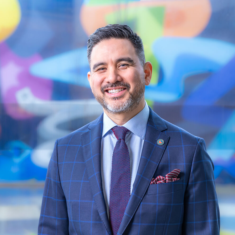 The image shows a smiling man in a suit and tie, likely a professional or politician, posing in front of a colorful background.