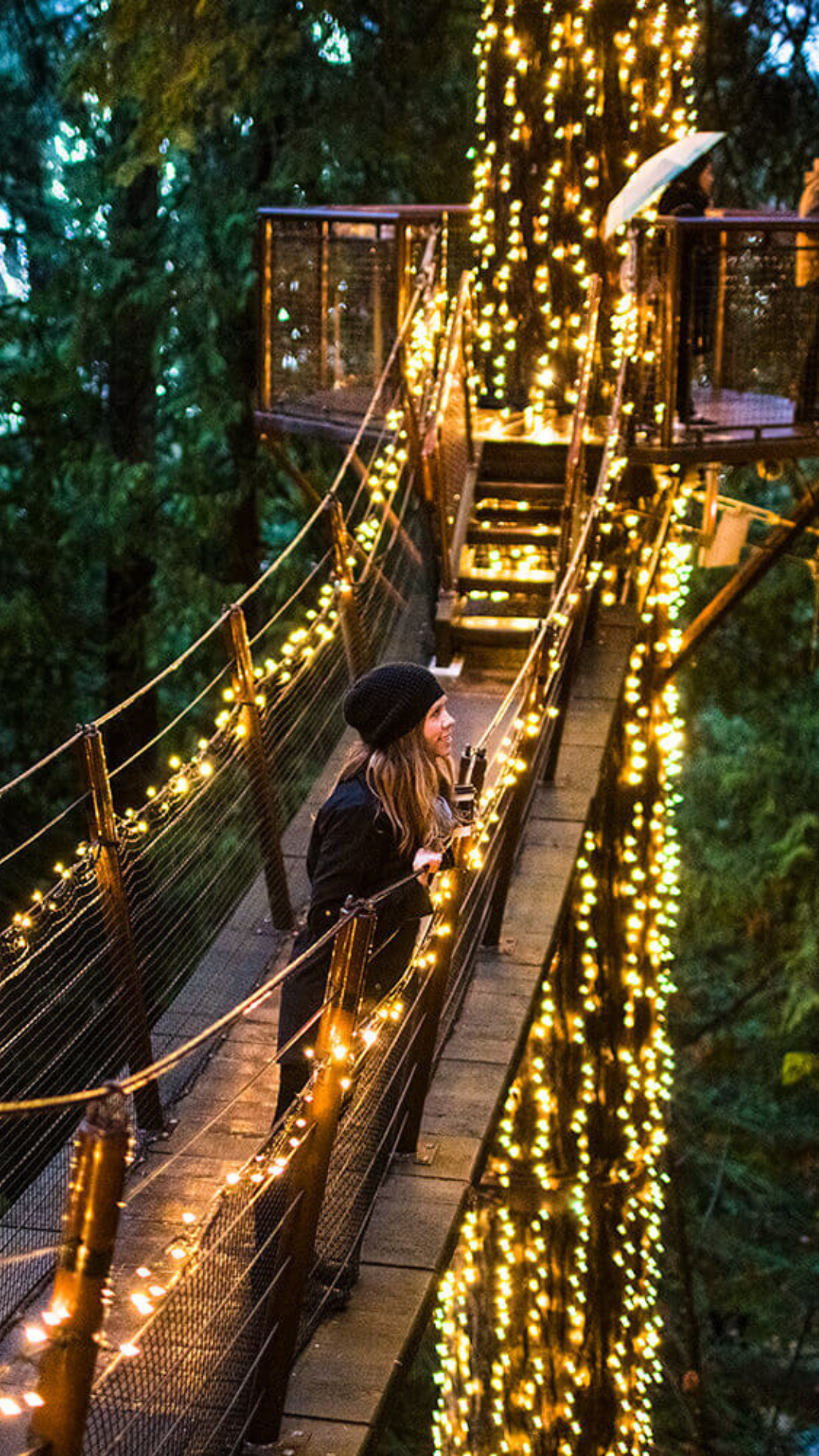 A woman enjoys a magical evening on a beautifully lit suspension bridge surrounded by dense forest.