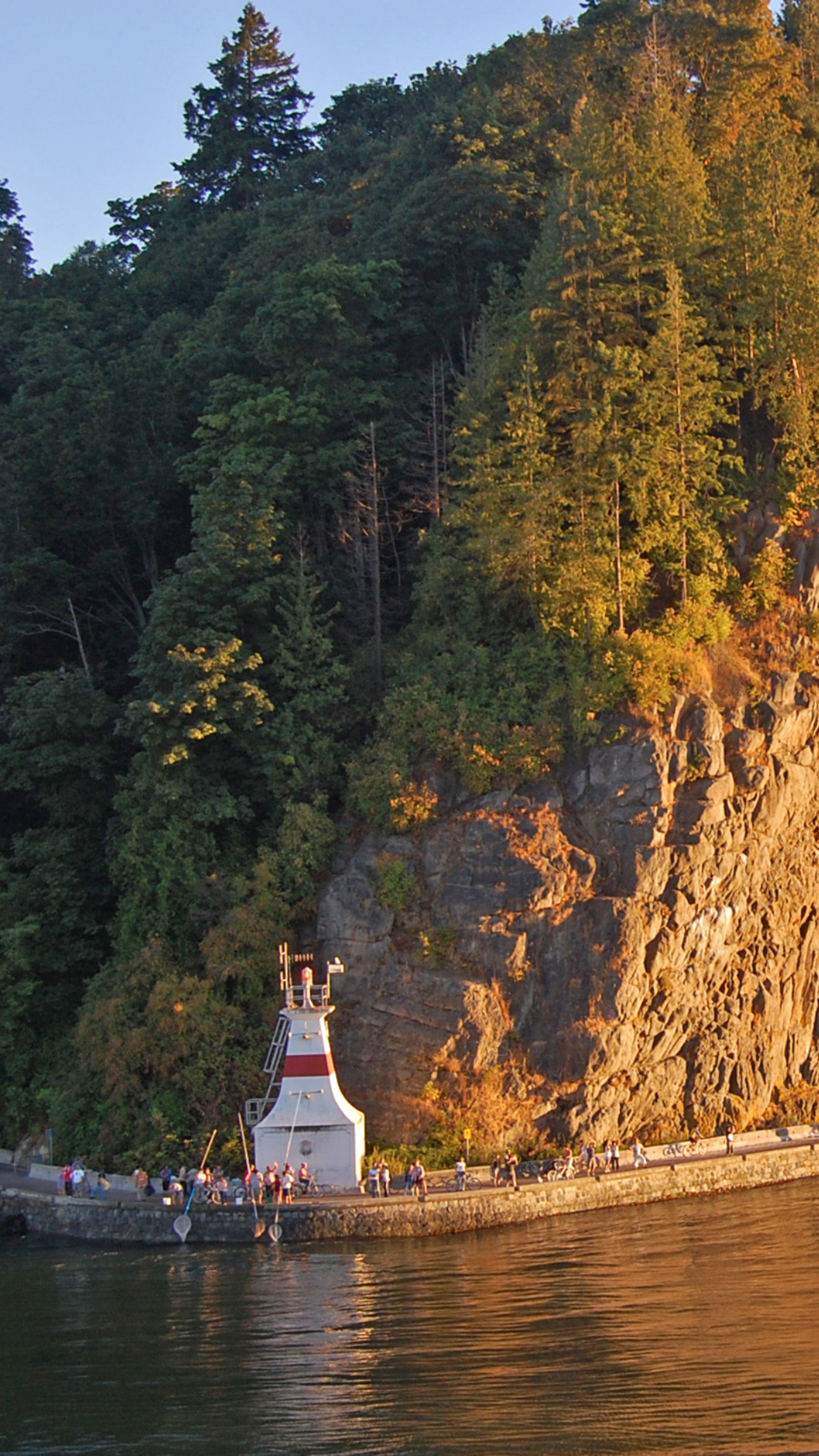 Une scène pittoresque montrant un phare sur une côte rocheuse entourée de forêts luxuriantes, baignée dans la lueur chaleureuse du coucher de soleil.