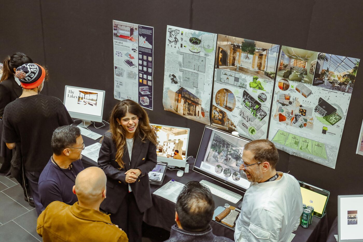 A group of people are gathered around a table displaying architectural designs and computer screens. A woman in a blazer appears to be presenting or explaining the designs to the others, who are listening attentively.