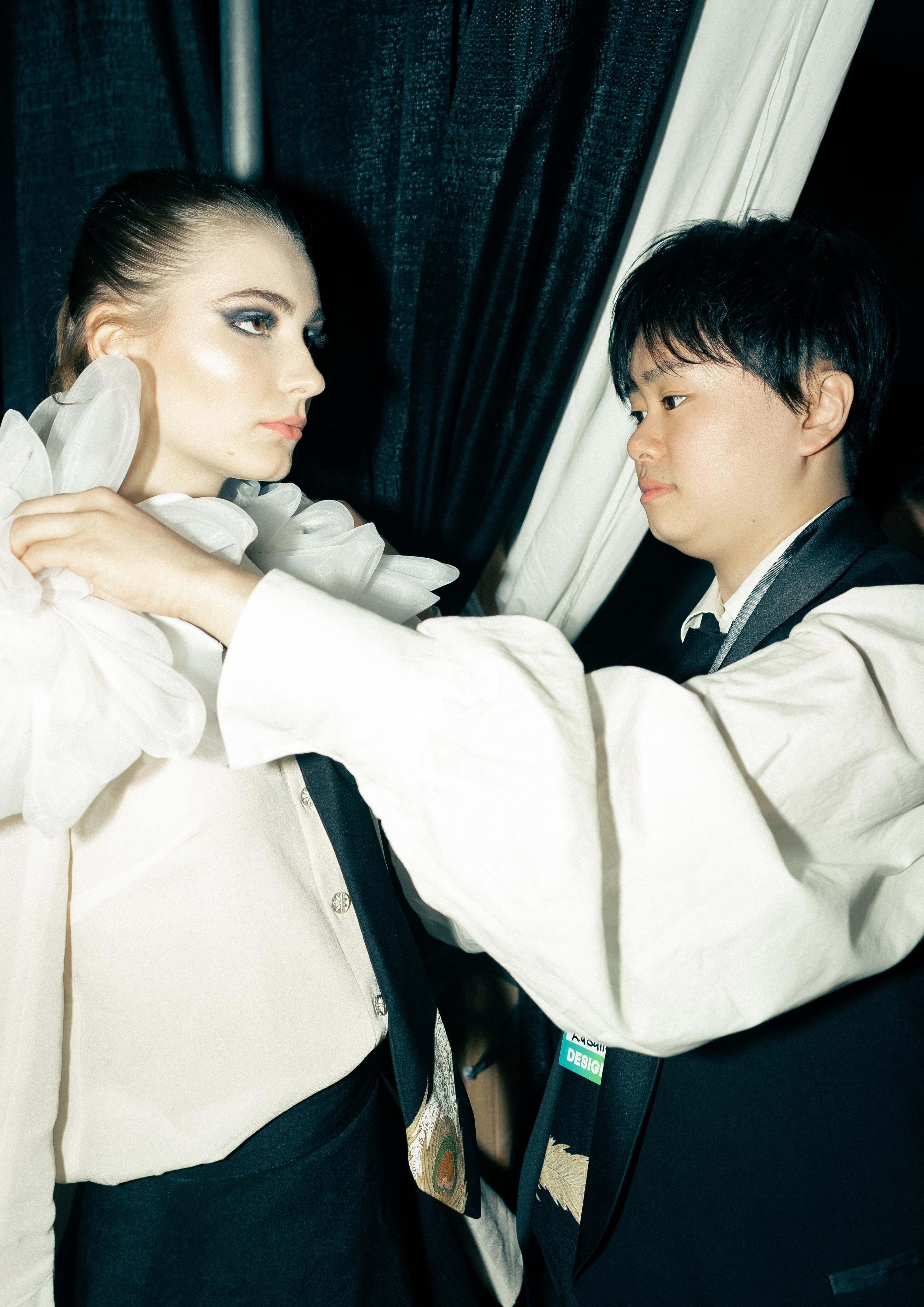 A young woman in a white shirt and puffy white collar is being helped by a person dressed in black, adjusting the collar for her.