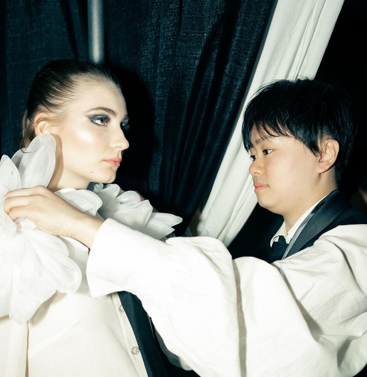 A young woman in a white shirt and puffy white collar is being helped by a person dressed in black, adjusting the collar for her.