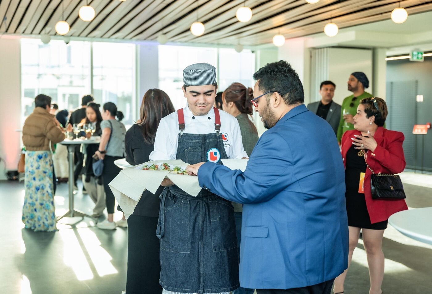 A chef in a white uniform presents a platter of food to a man in a blue blazer, surrounded by other people at a catered event.