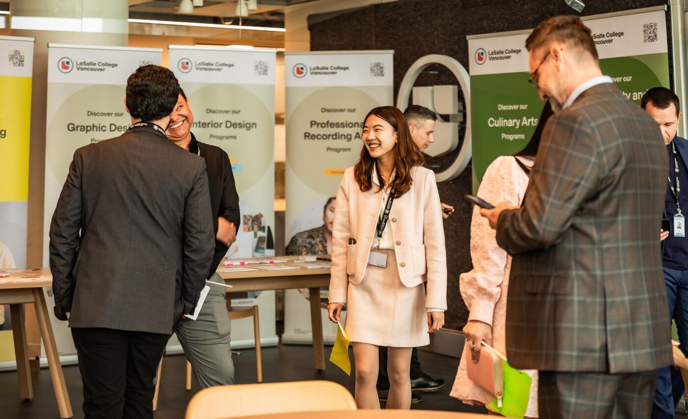 A group of people stand in a room with various college program posters displayed on the walls, some people are interacting while others look at their phones.
