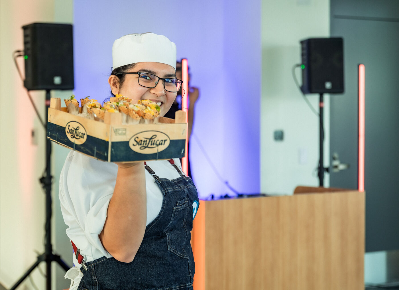 A woman wearing a chef's hat and apron holds a wooden crate filled with food, smiling at the camera in a room with speakers and a light-purple background.