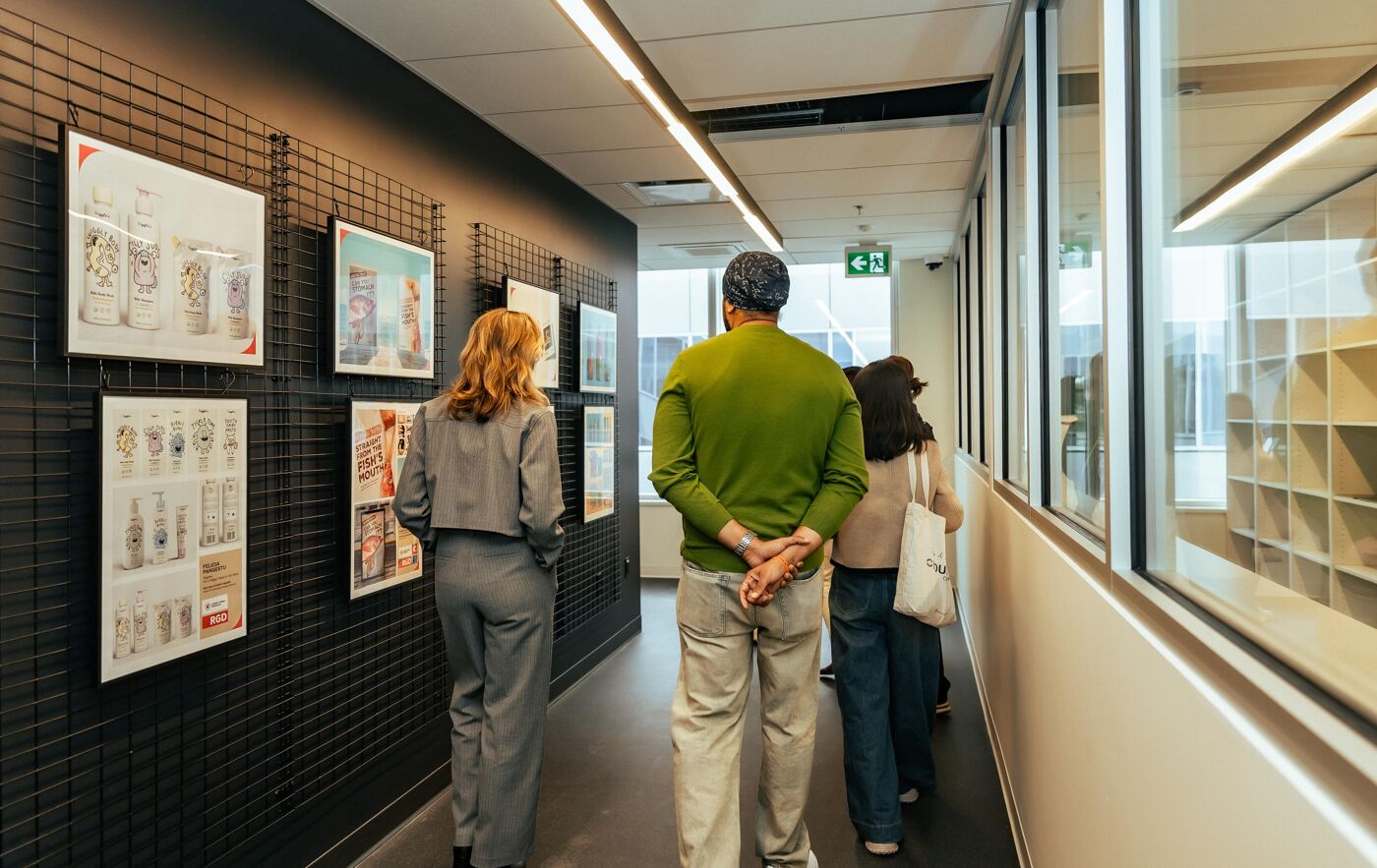 A group of people stand in a hallway viewing artwork displayed on a black grid wall, with large windows showing an exterior view on the right.