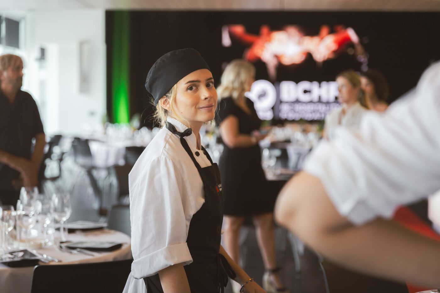 A young female chef wearing a cap and black apron looks towards the camera at an event with set tables and other people in the background.