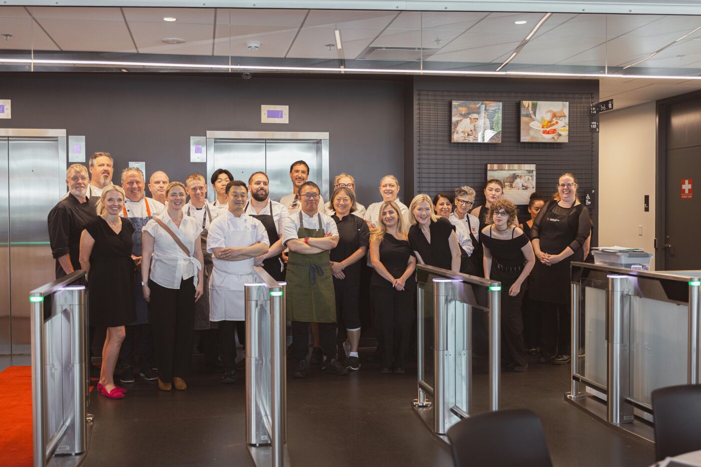A group of chefs and front-of-house staff pose together in front of elevators and turnstiles, dressed in black and green uniforms.