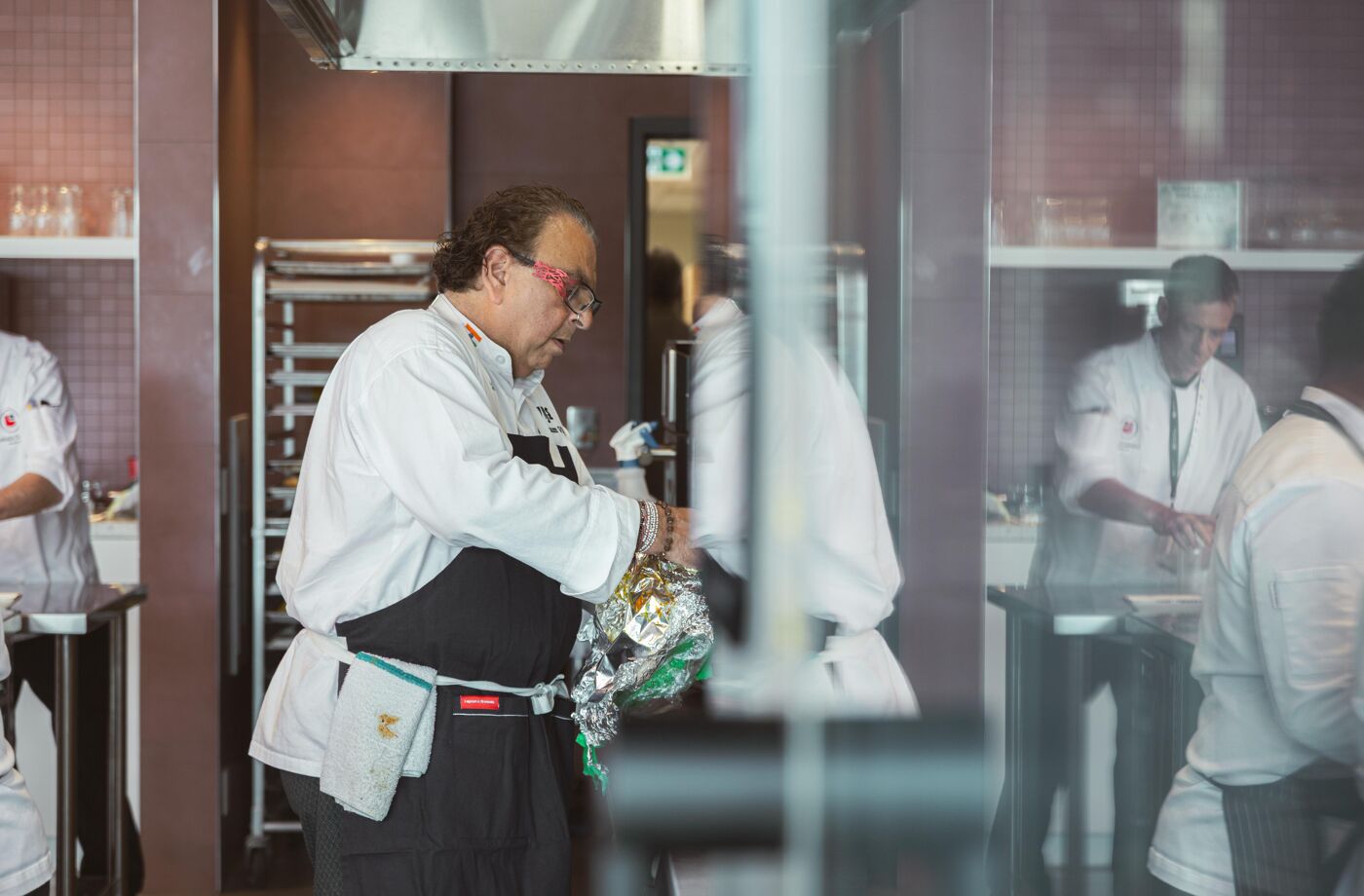 A chef wearing glasses prepares ingredients wrapped in aluminum foil in a professional kitchen, surrounded by other cooks working on their own dishes.