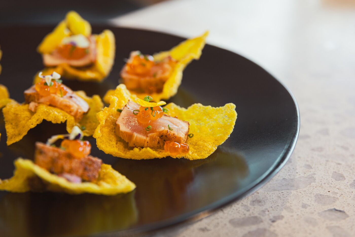 These are salmon tartare bites with roe eggs on yellow wonton crisps, presented on a black plate. They are an elegant and flavorful appetizer.