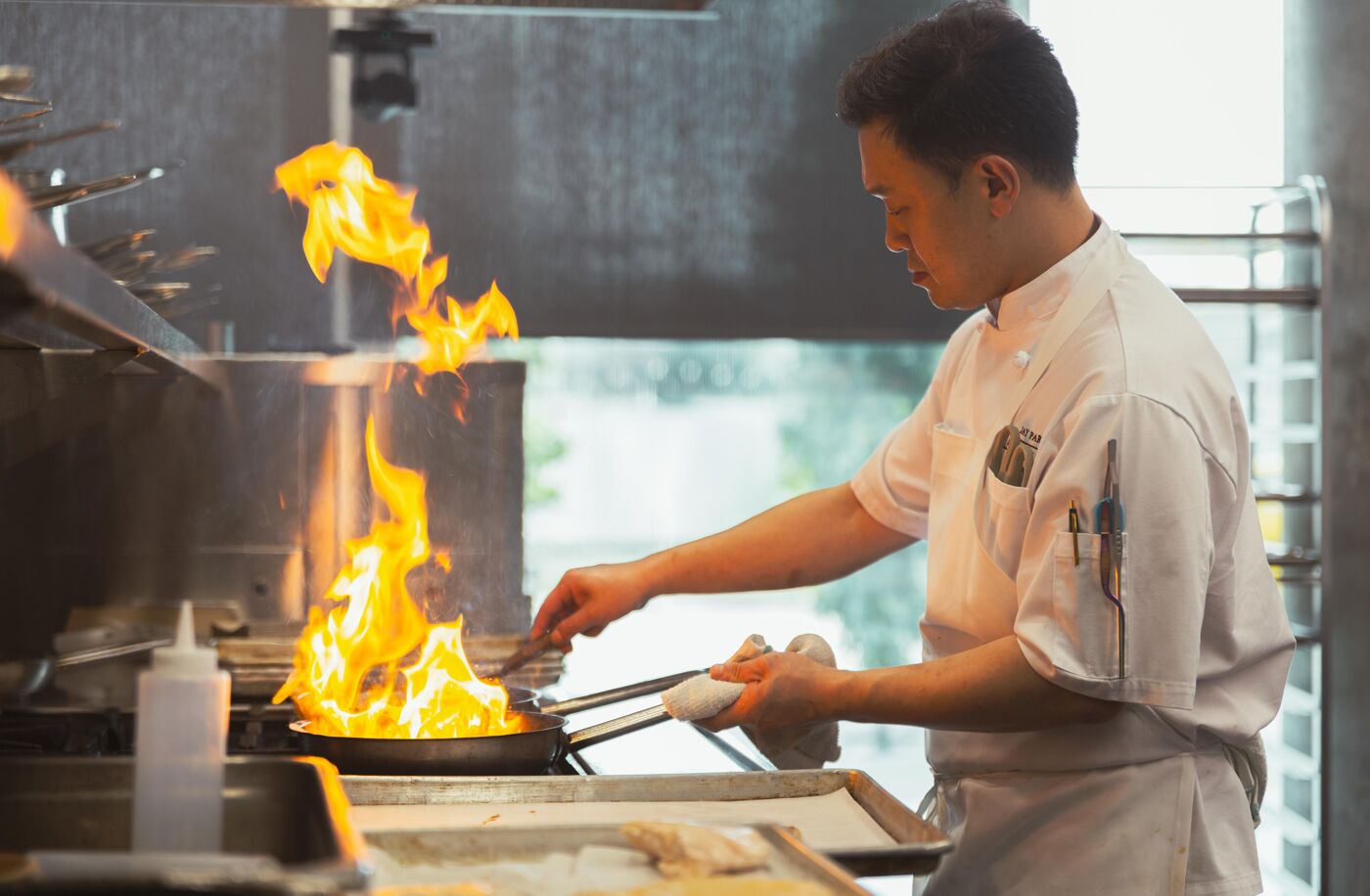 A professional chef is cooking with open flames in a pan, creating a culinary spectacle with vibrant fire in a professional kitchen setting.