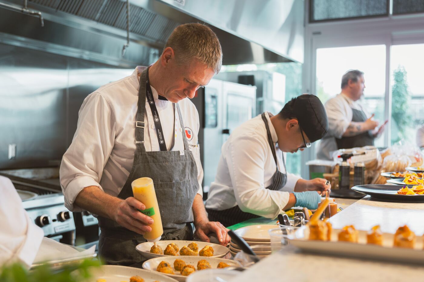 Chefs in a professional kitchen are carefully plating small appetizers, with one chef artfully drizzling sauce from a squeeze bottle onto the dishes.