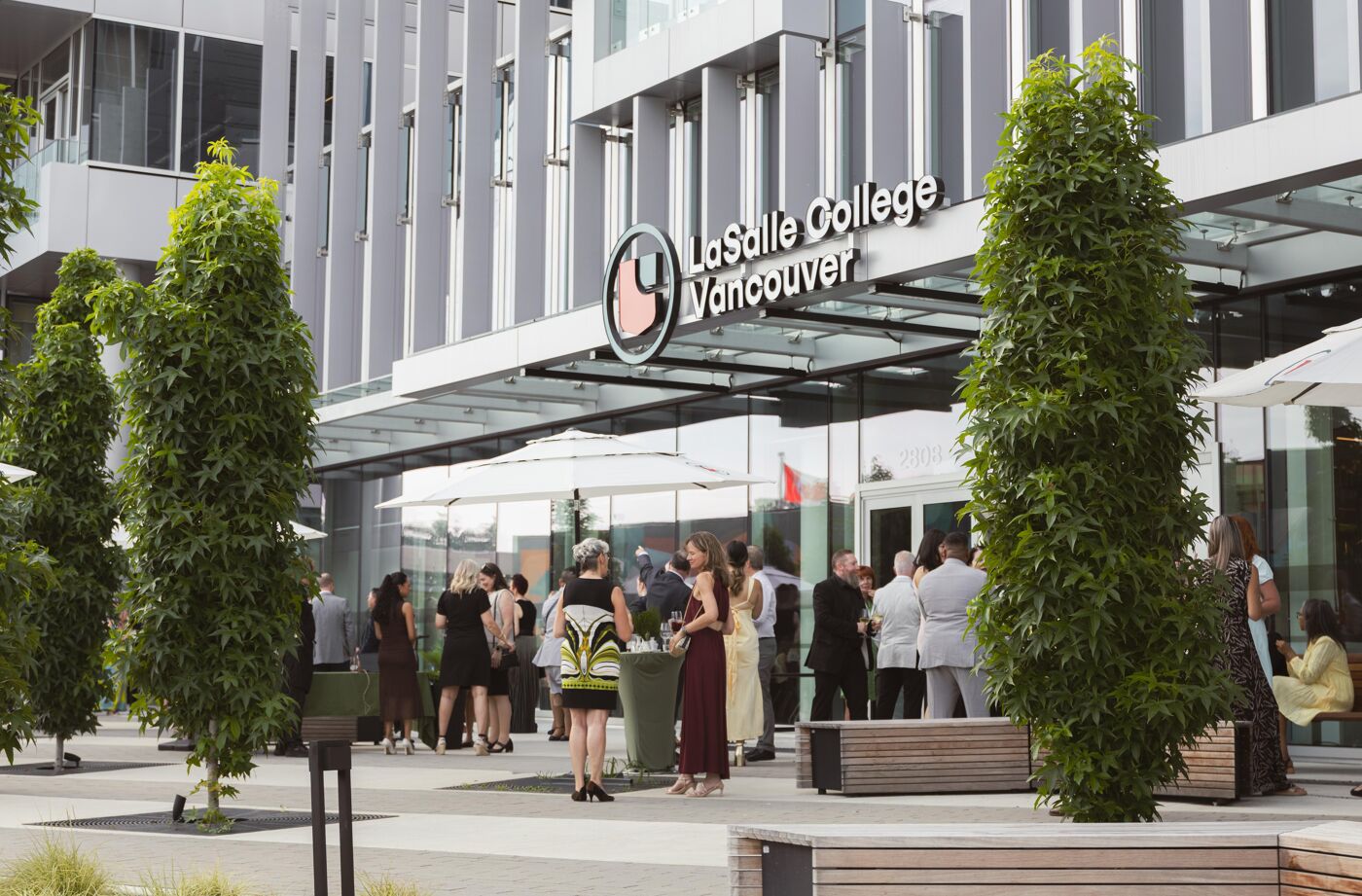 A group of people are gathered outside the LaSalle College Vancouver building, enjoying an outdoor event with green trees and a sunny day.