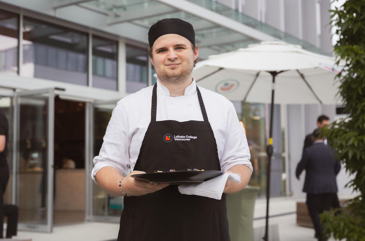 A smiling chef holds a plate of desserts in front of a modern building. He is wearing a black chef's hat and a black apron over a white uniform.