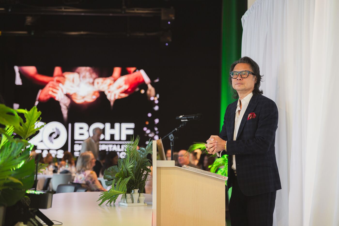 A man wearing glasses and a suit stands at a podium, speaking in front of a screen displaying the "BCHF 20 YEARS" logo.