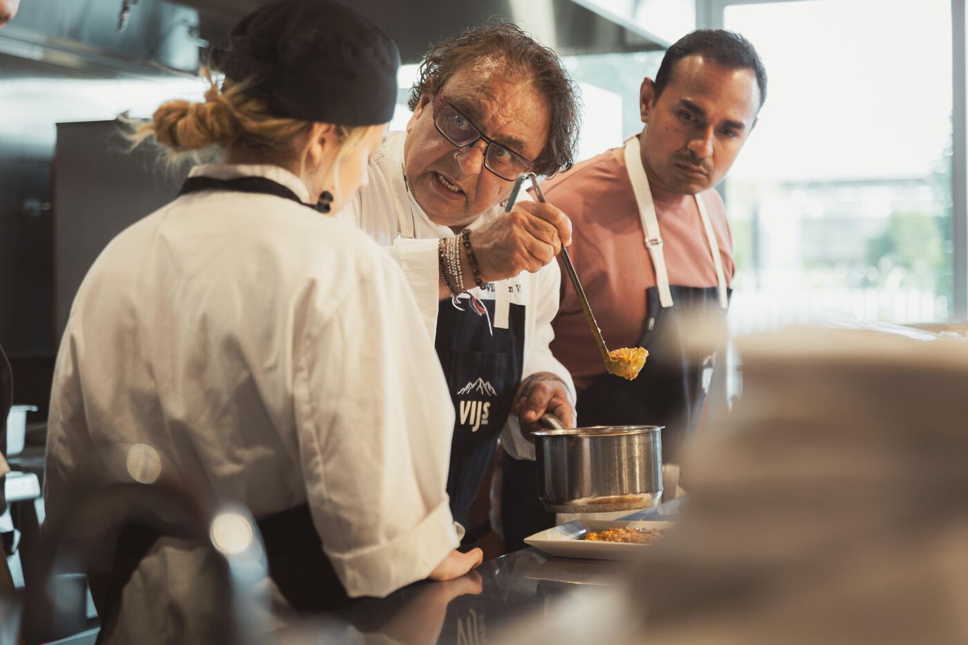 An experienced chef wearing glasses and a black apron is instructing two cooks in a professional kitchen, showing an ingredient from a pot.