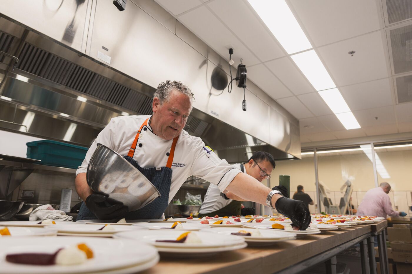 Chefs in a commercial kitchen are meticulously arranging food on plates, preparing for a large event or service. The focus is on detailed presentation and efficient workflow.