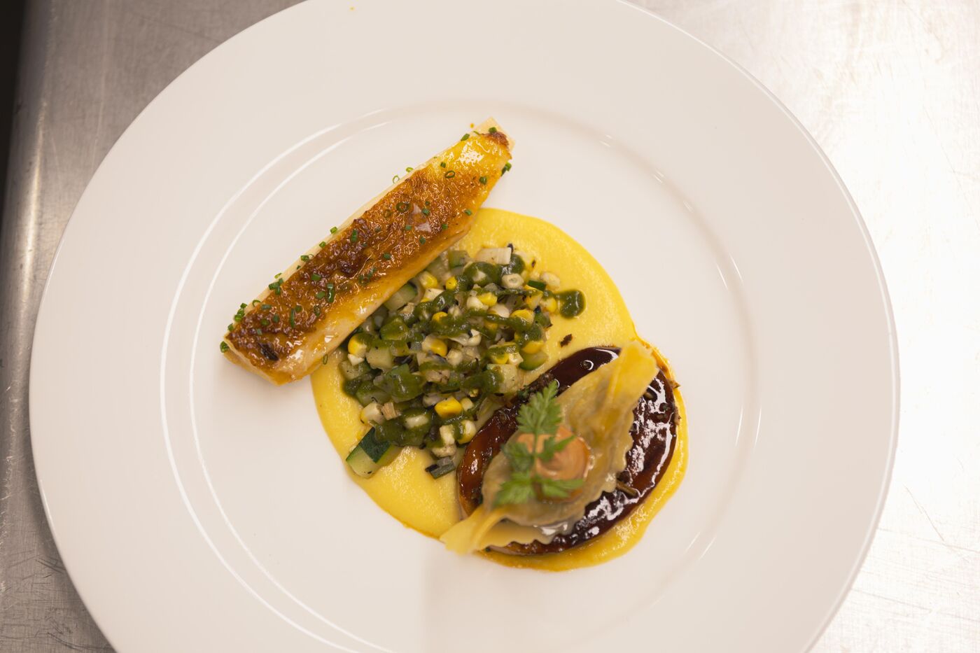 A gourmet dish featuring crispy fish, corn puree, chopped vegetables, and a dark sauce, artfully presented on a white plate.