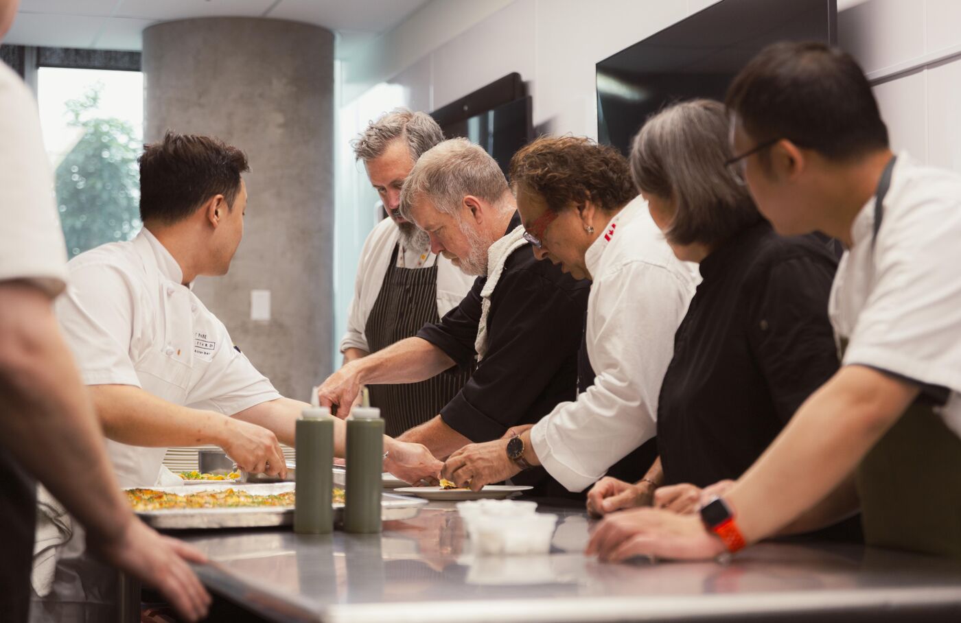 A group of chefs are working together in a kitchen, preparing food at a workstation. It appears to be a cooking class or culinary demonstration.