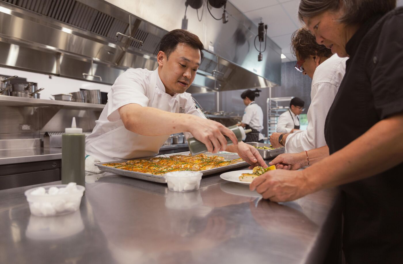 An Asian chef is adding a final touch to a dish in a professional kitchen, with other cooks working in the background. The food looks delicious and carefully prepared.
