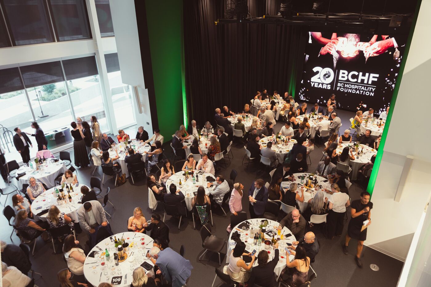 Aerial view of a gala event with guests seated at round tables, featuring a large illuminated "20 Years BCHF Hospitality Foundation" logo on screen.