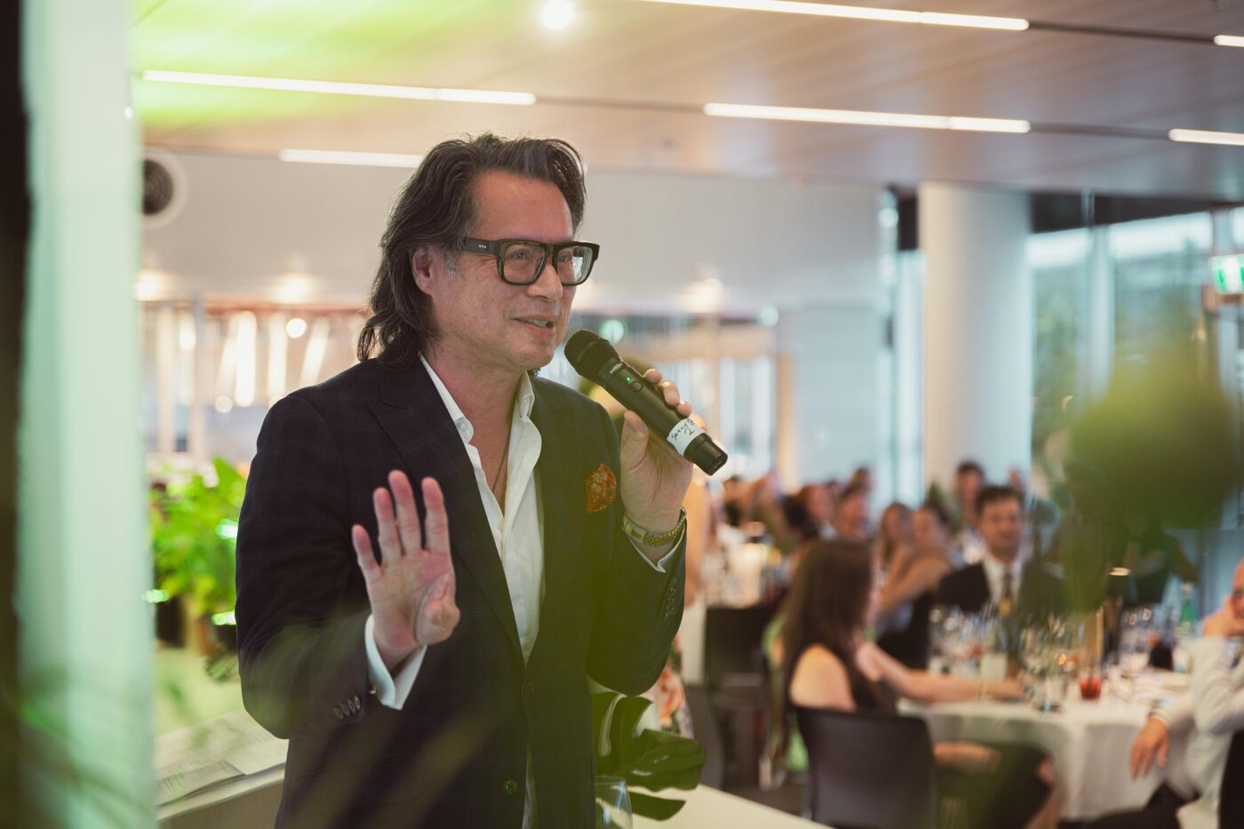 A man wearing glasses and a suit is speaking into a microphone in front of an audience seated at tables.