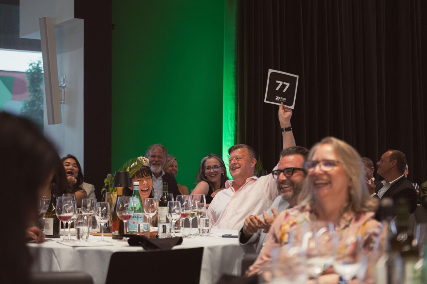 A group of people are laughing and enjoying an event, possibly an auction, with a man holding up a sign with the number 77.