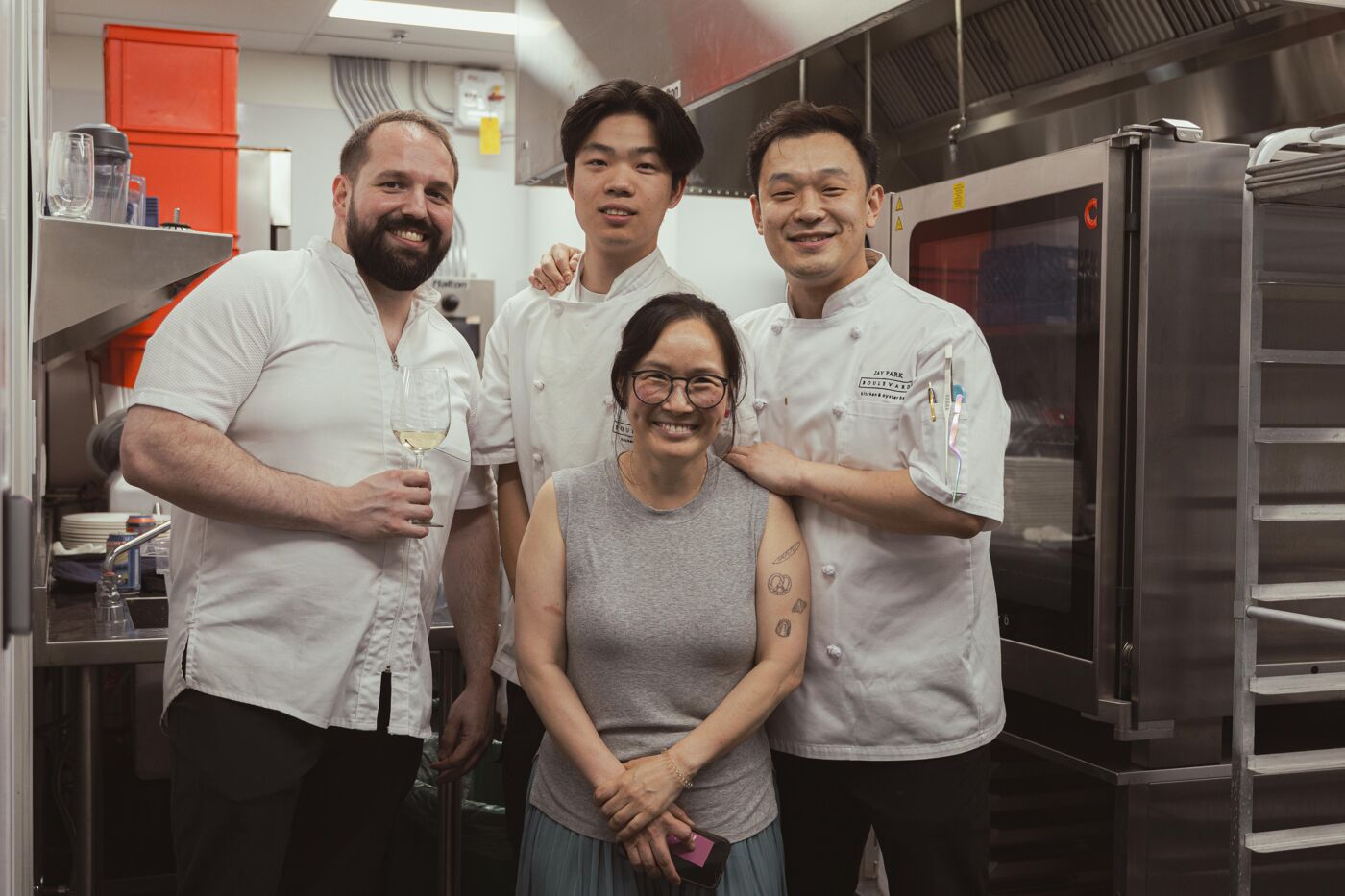 Four people smile at the camera in a professional kitchen. Two men with beards wear white chef hats, another man in a white chef hat and a woman wearing glasses stand in front.