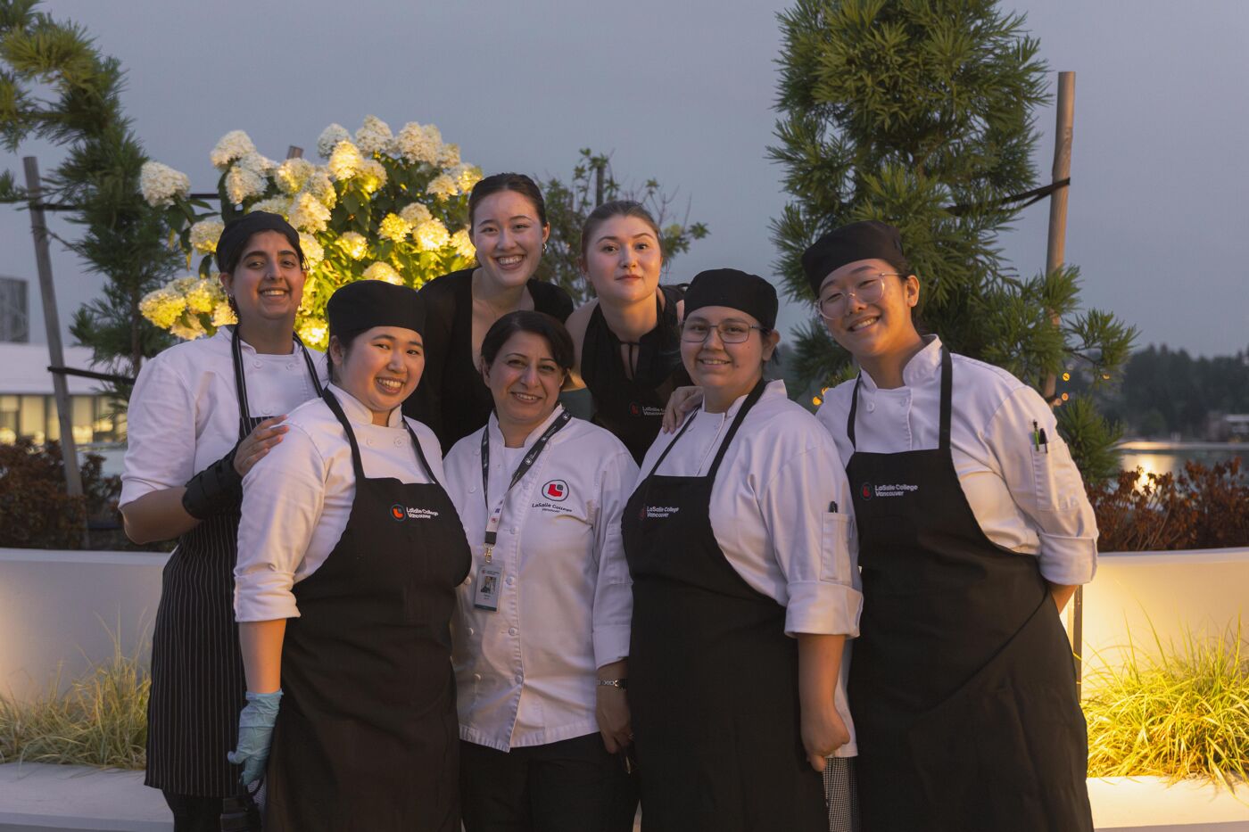 A group of seven smiling chefs, mostly women, pose for a photo in an outdoor setting with flowers and greenery.