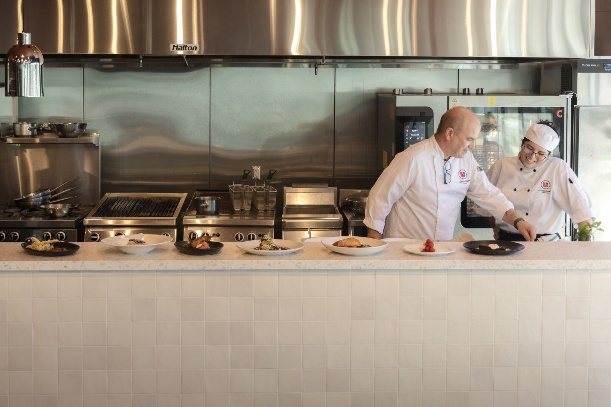 Two chefs are in a kitchen, examining plates of food on a counter. The kitchen has stainless steel appliances and a clean, modern design.