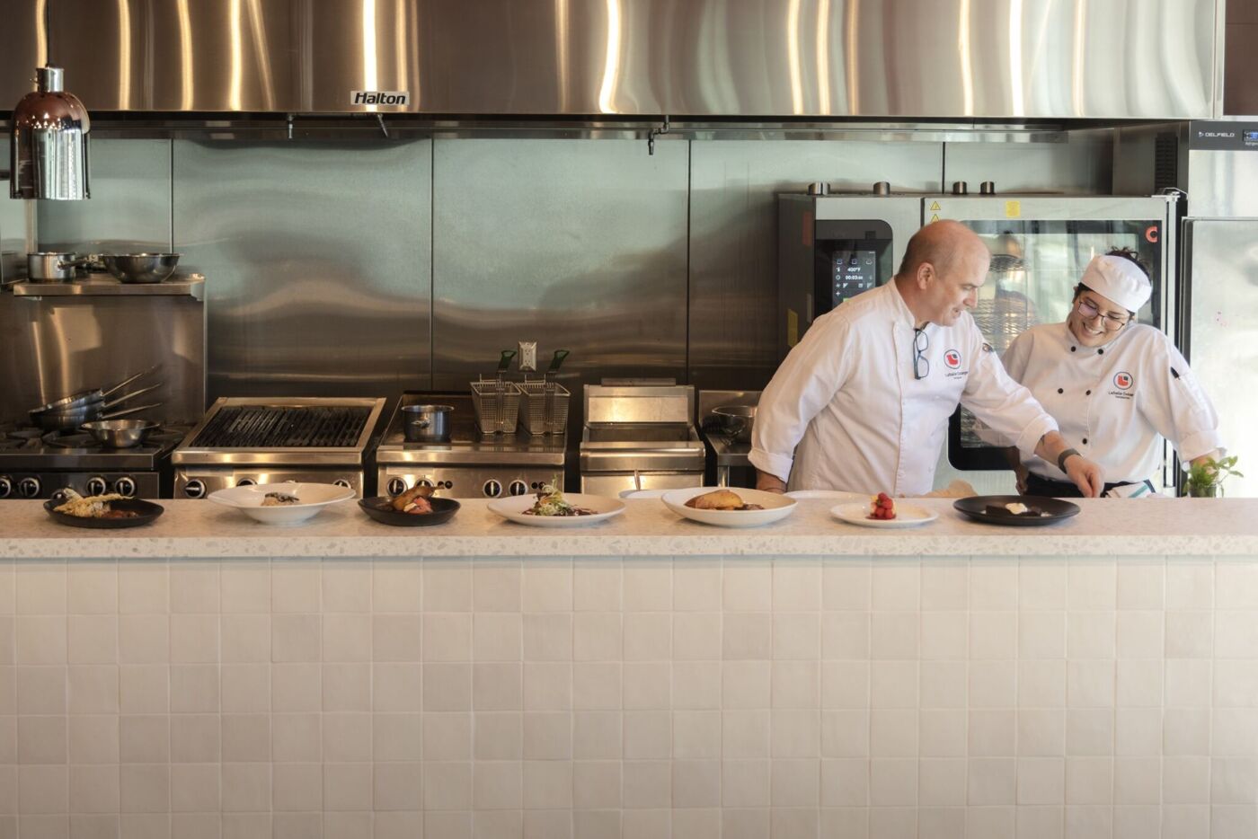 Two chefs are in a kitchen, examining plates of food on a counter. The kitchen has stainless steel appliances and a clean, modern design.