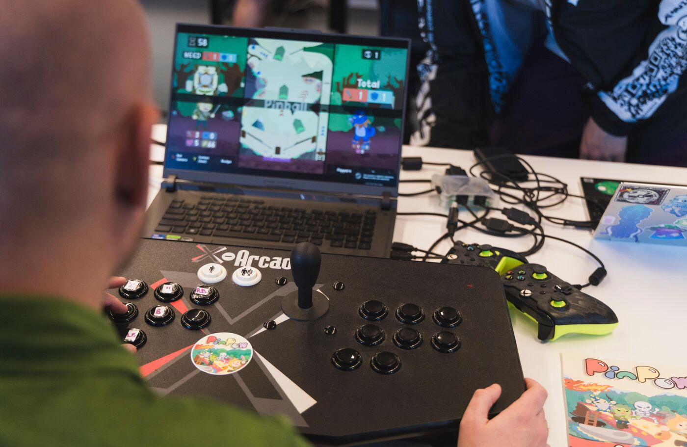A person plays an arcade fighting game connected to a laptop, with a joystick and another controller to the right.