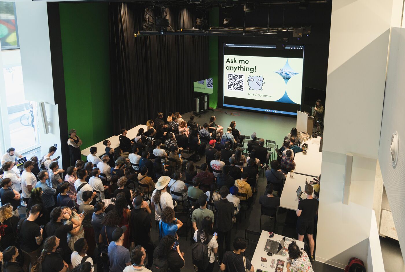 A large audience is attending a presentation with a screen displaying "Ask me anything!" along with QR codes.