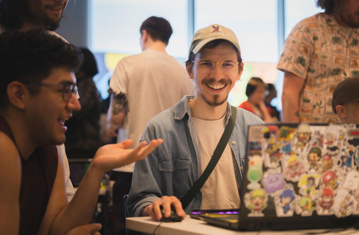 A smiling man wearing a cap and glasses, seated in front of a sticker-covered laptop, interacts with another man.