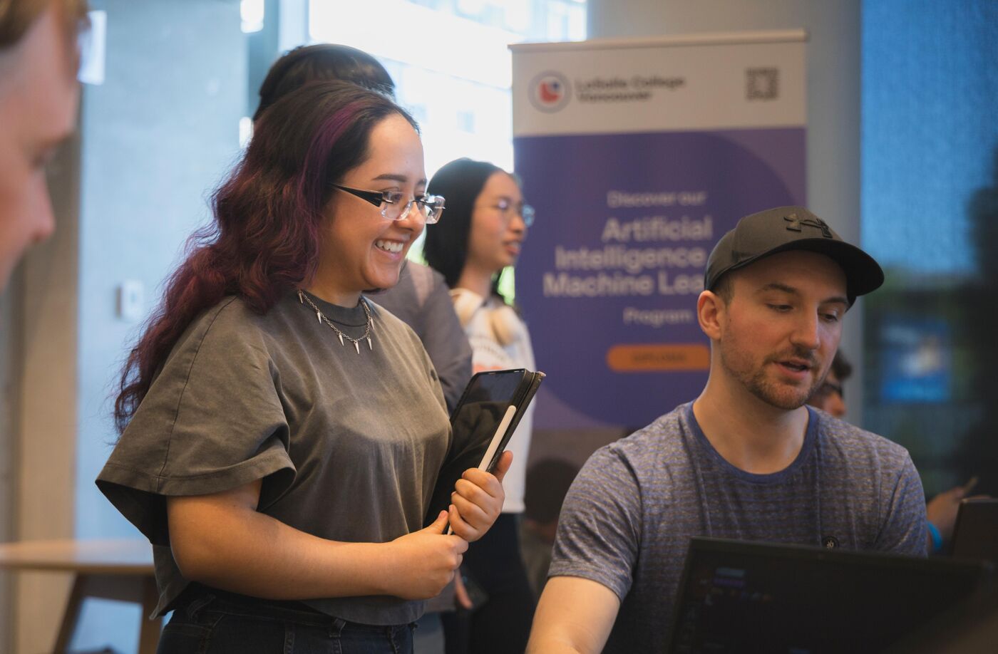 A man and a woman smile while looking at a computer screen, with other people in the background near a banner about artificial intelligence and machine learning.