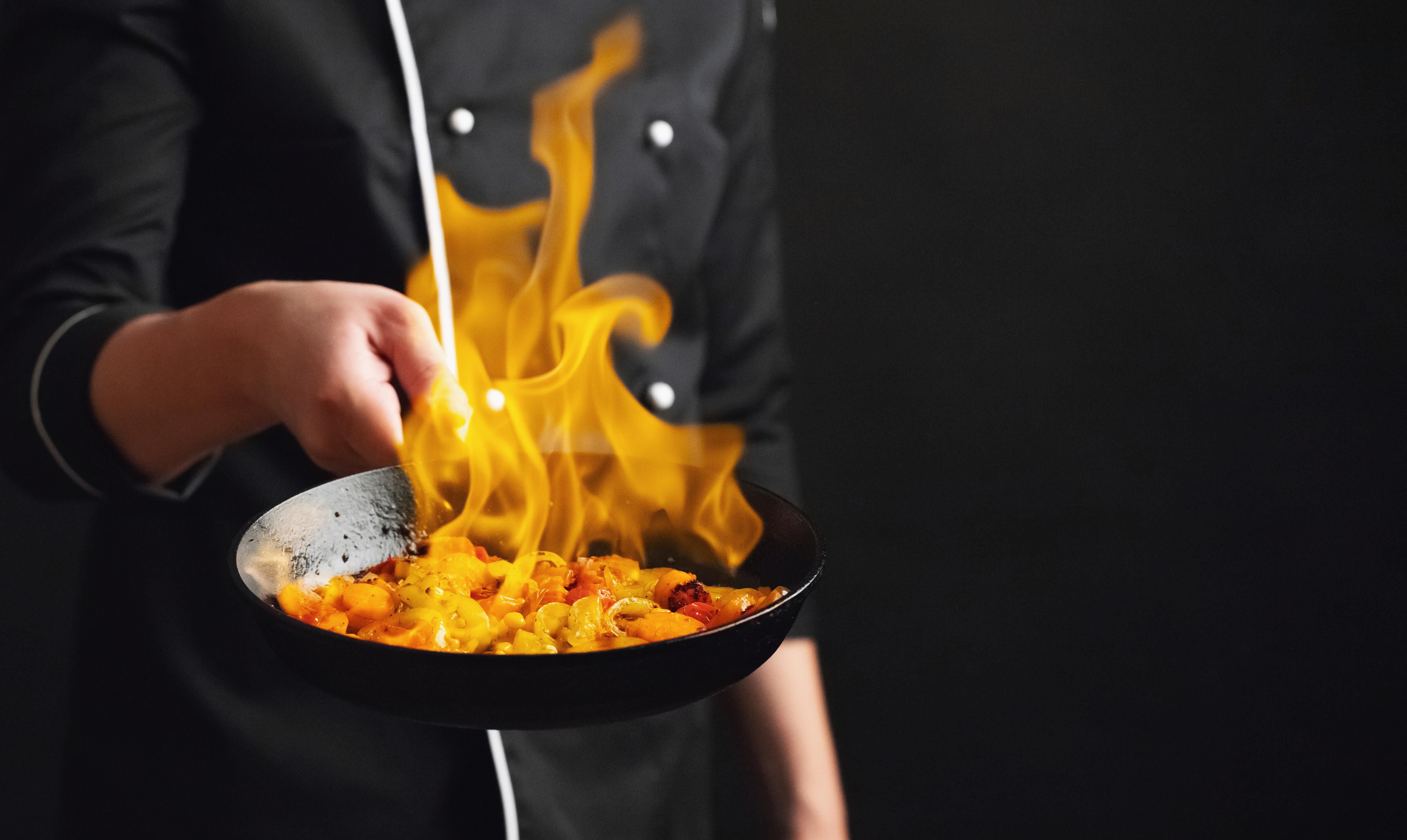 A chef in a black uniform is holding a pan with food on fire, creating a dramatic culinary scene against a dark background.