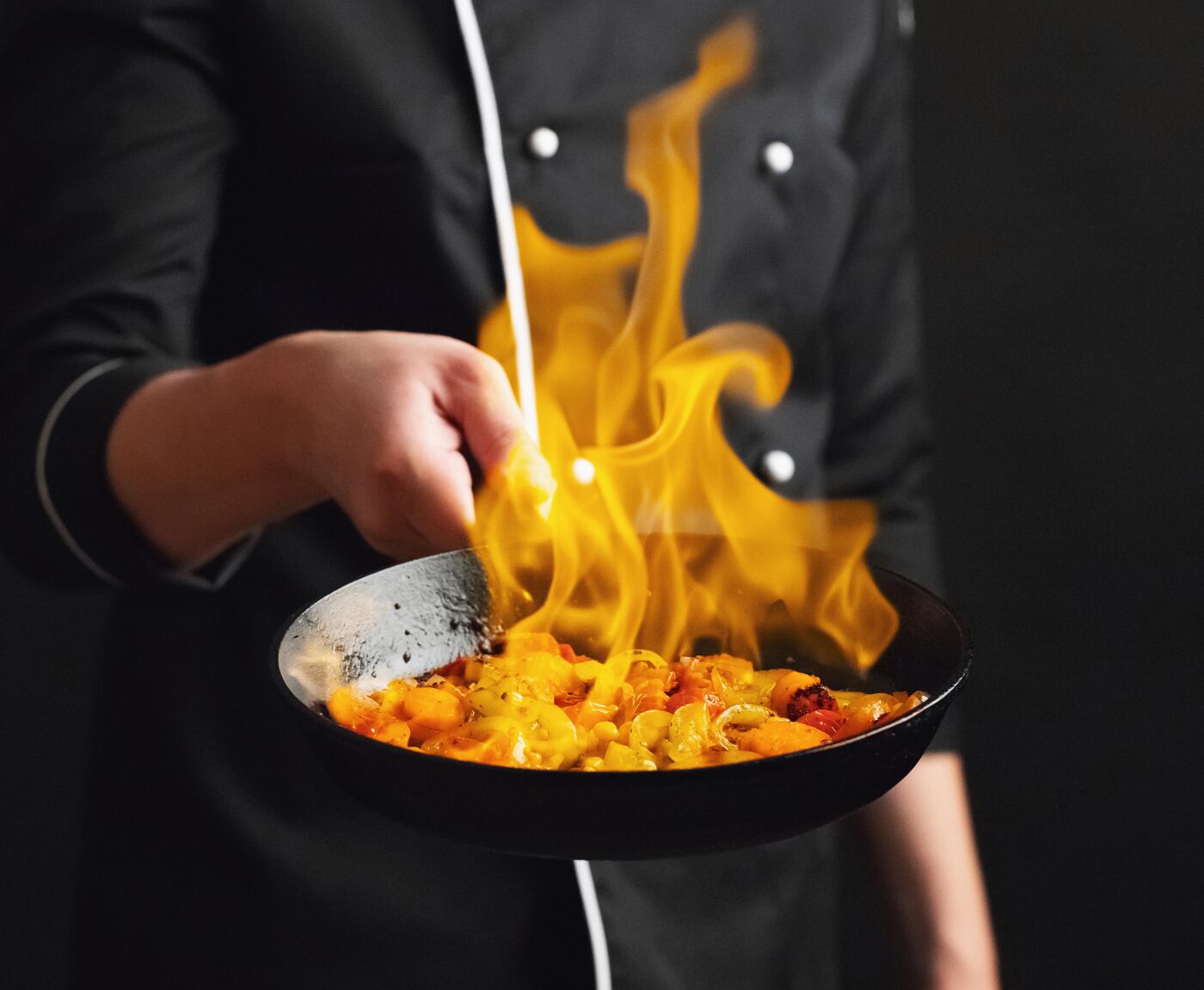 A chef in a black uniform is holding a pan with food on fire, creating a dramatic culinary scene against a dark background.