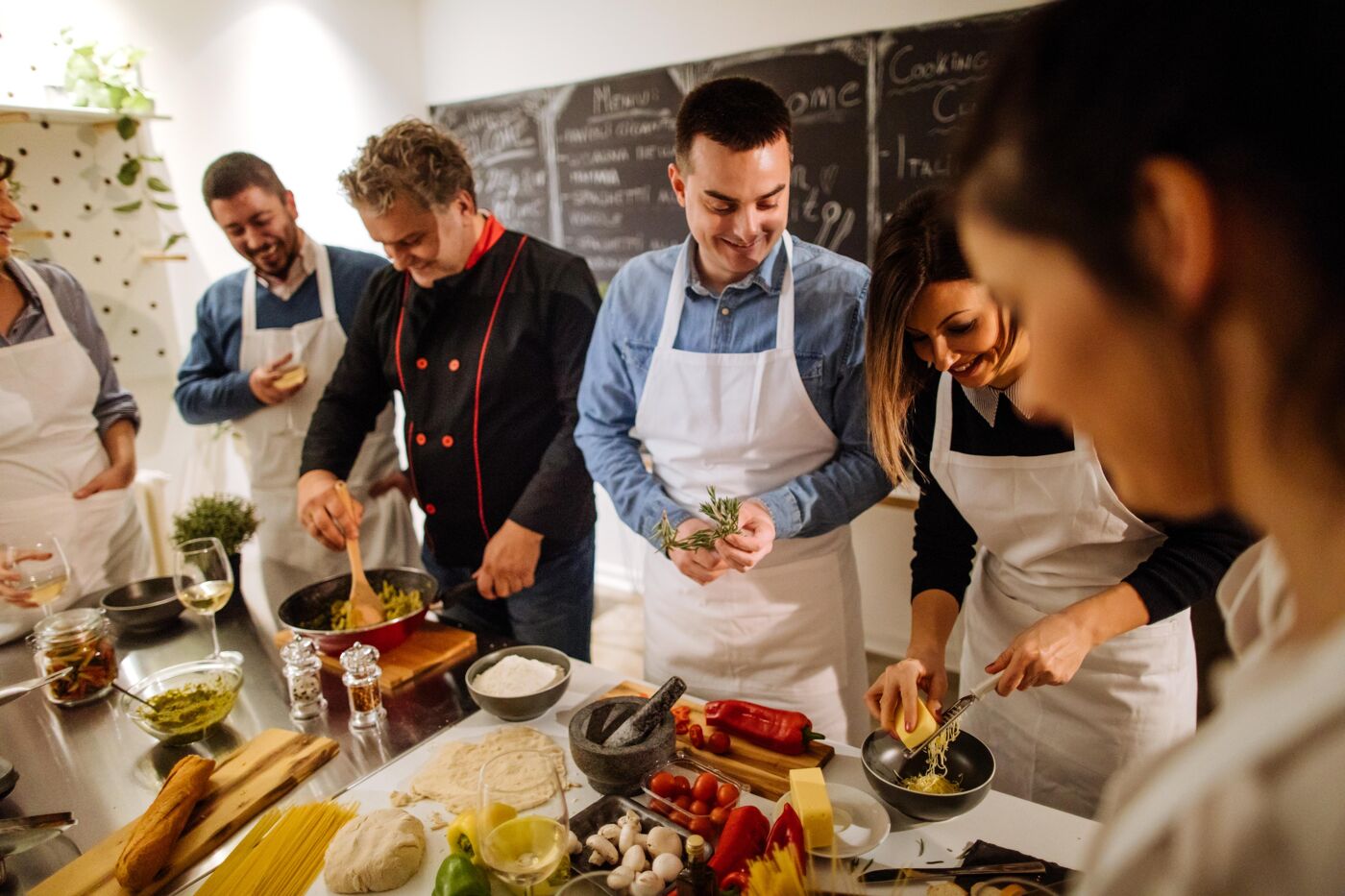 The image shows a cooking class with several people preparing food together. They are wearing aprons and seem to be enjoying the process of cooking and learning.