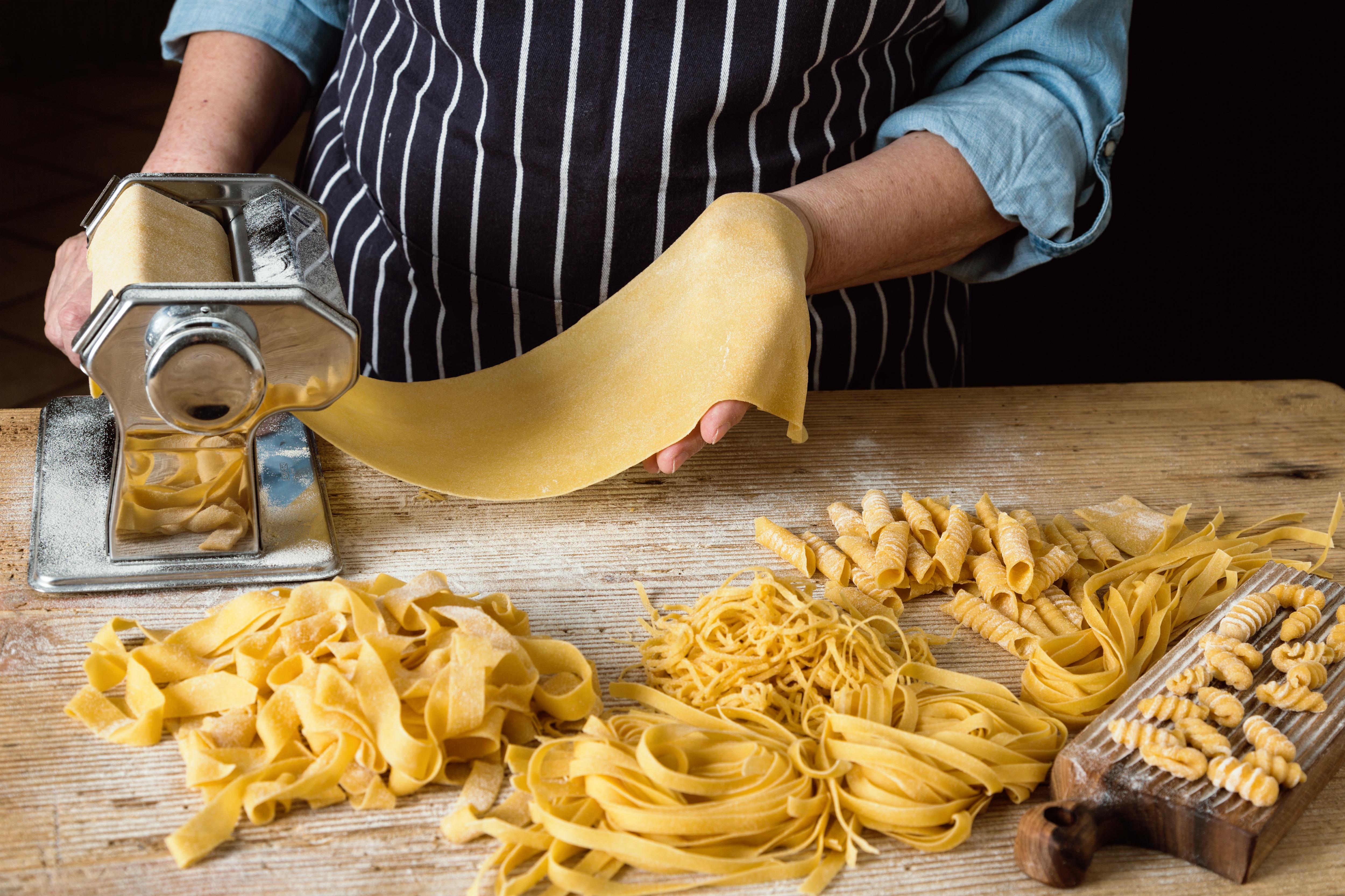The image shows a person making pasta with a pasta machine. There are different types of pasta on a wooden table, including fettuccine and fusilli.