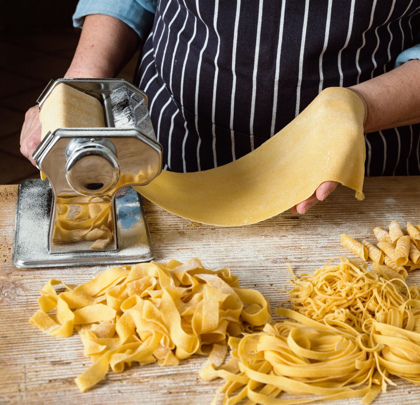 The image shows a person making pasta with a pasta machine. There are different types of pasta on a wooden table, including fettuccine and fusilli.