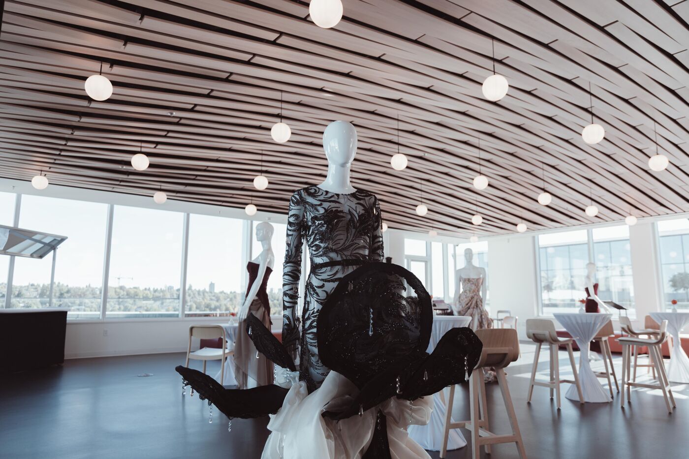 The image shows a fashion exhibition with mannequins displaying avant-garde outfits. The room features a modern design with a slatted ceiling and large windows, creating a bright and airy atmosphere.