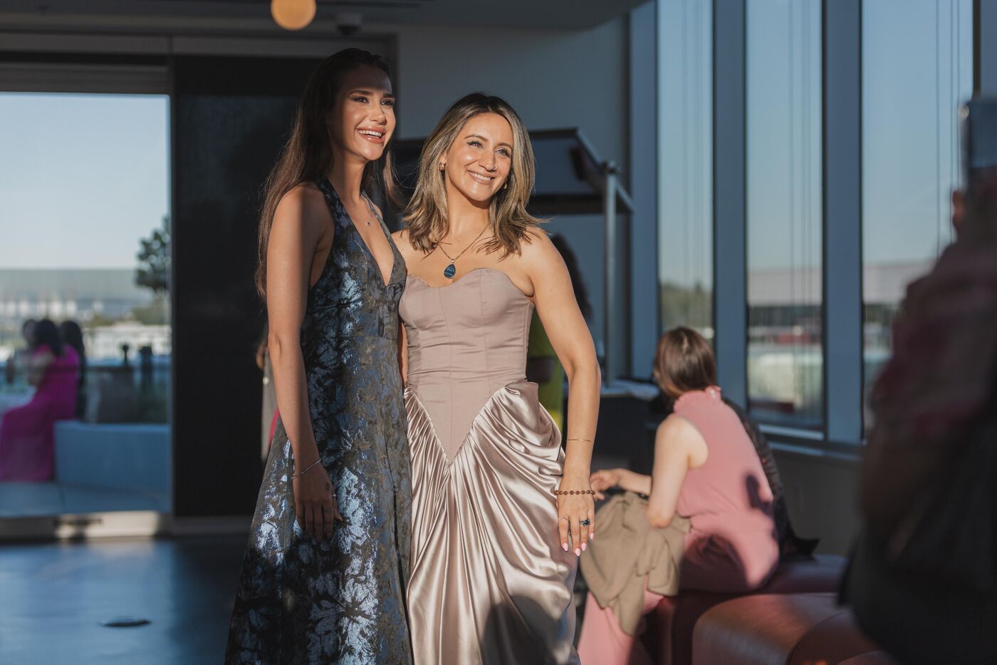 Two women in elegant dresses pose for a photo indoors, with large windows offering a view of the outdoors. They are smiling and appear to be at a formal event.
