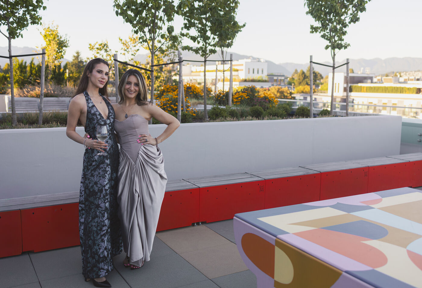 Two women in elegant dresses pose for a photo on a rooftop patio with a cityscape in the background. The woman on the left wears a sequined gown, while the woman on the right is in a draped, satin dress.