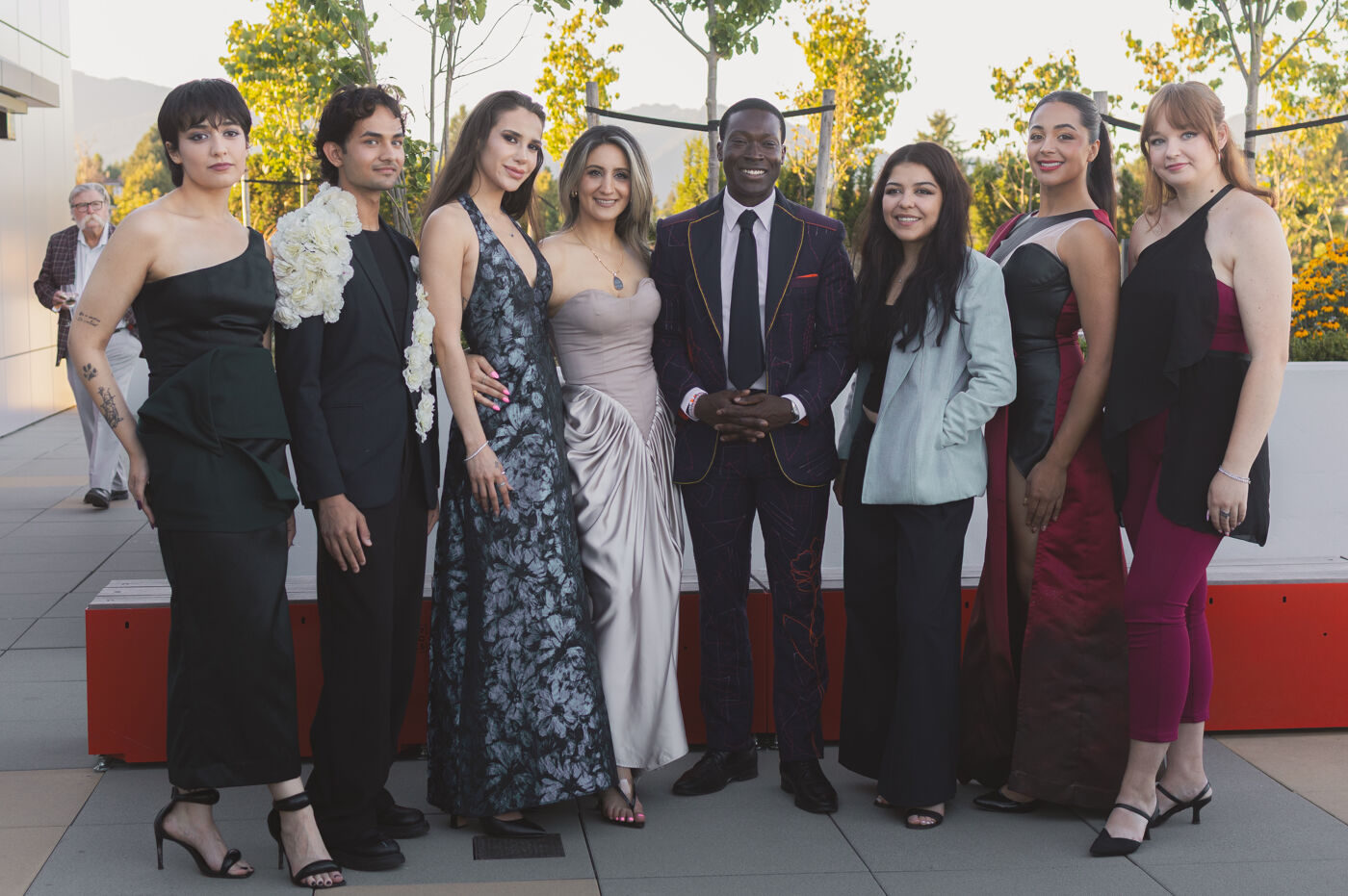 A diverse group of nine people, dressed in formal attire, pose for a photo outdoors. The group includes six women and three men, with varying ethnicities and styles.