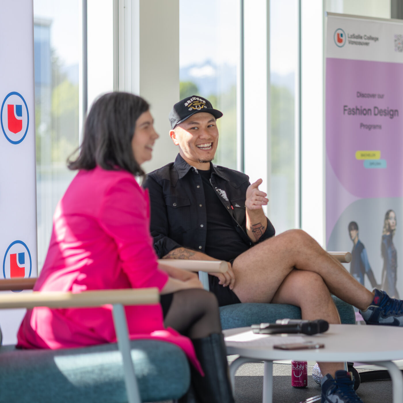 Two people are seated in chairs, talking at an event. The woman is wearing a fuchsia jacket and the man is wearing a cap and a black t-shirt.