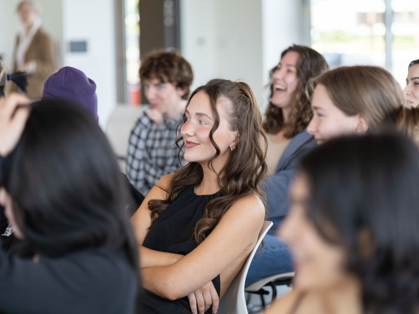 A group of young people are attending a presentation or lecture, with some individuals laughing and others listening attentively. The atmosphere appears engaged and lively.