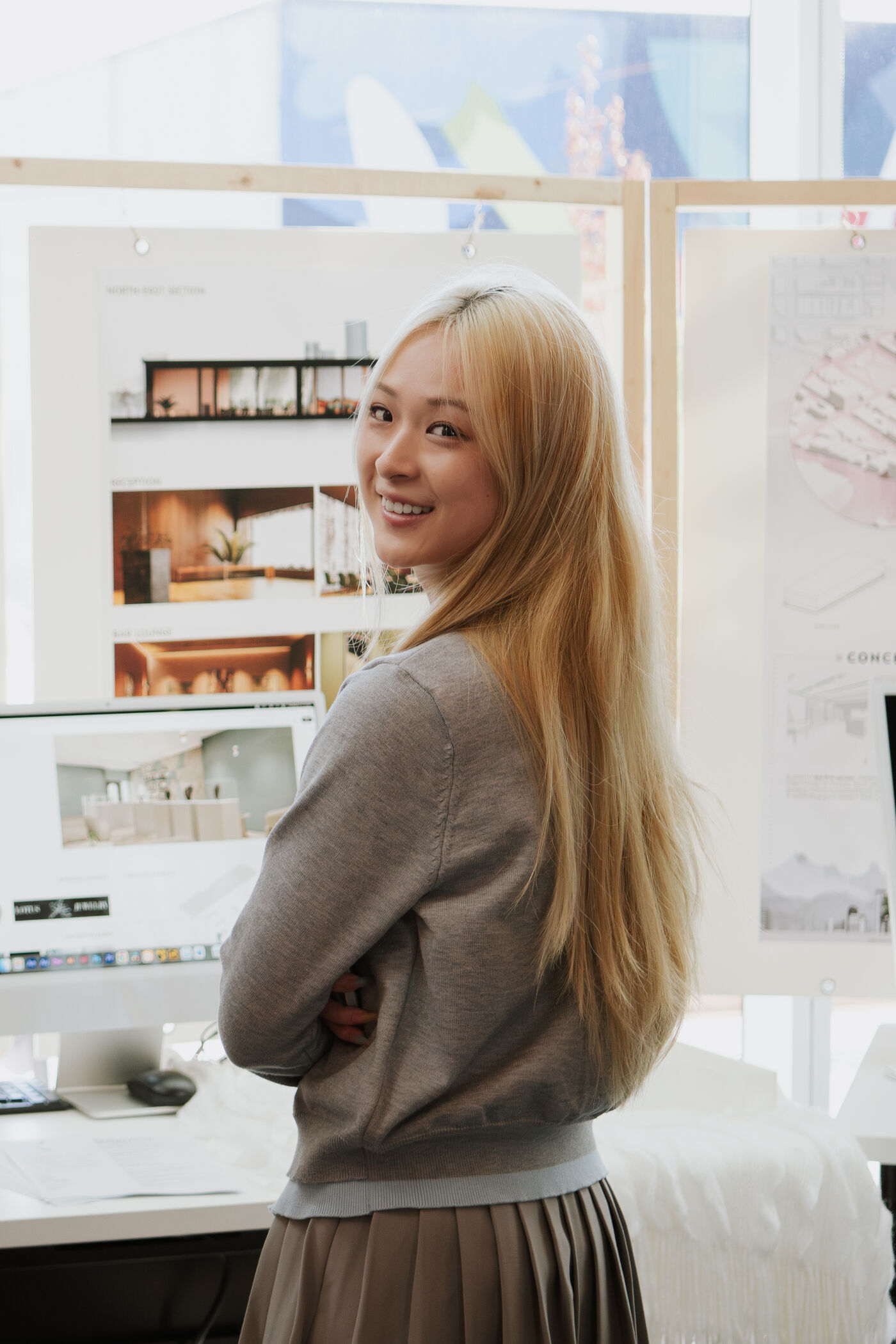 The image shows a young woman with long blonde hair, smiling and looking over her shoulder. She is standing in front of a desk with architectural plans.