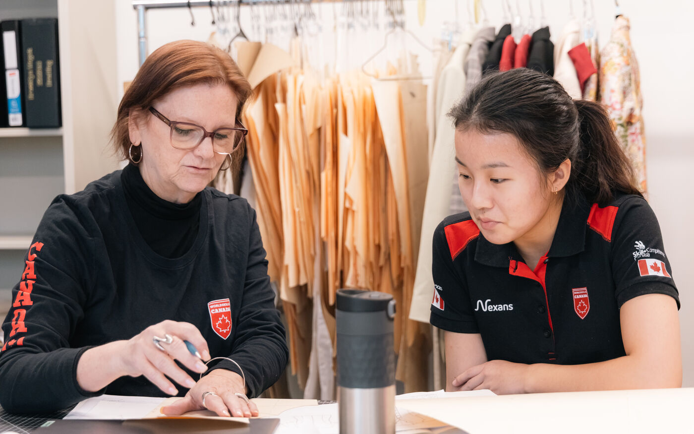 Two women, one older with glasses and a younger Asian woman, are looking at documents at a table, with clothes hanging in the background.