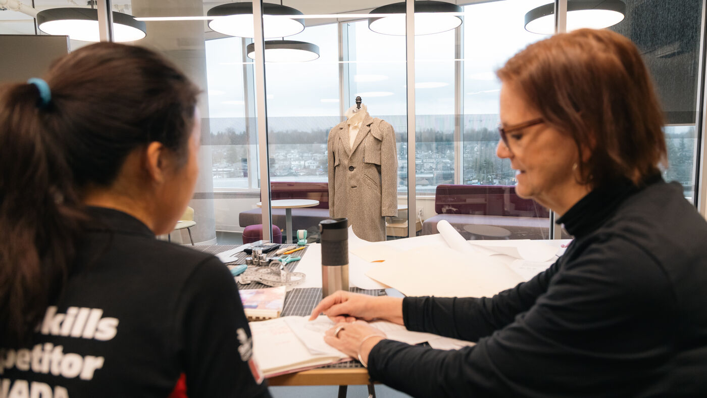Two women are seated at a table, reviewing documents. In the background, a fashion garment is displayed on a mannequin, with a large window offering outdoor views.